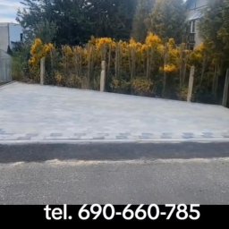 Brukisiema Mateusz Szczepaniak - Jasnoszara kostka brukowa tworząca podjazd, widoczna przez otwartą bramę, z roślinnością w tle. Estetyczne wykończenie posesji.