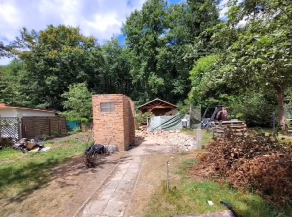 Budowa z cegły, widok na ogród z drzewami i altaną. W tle minikoparka i gruz, sugerujące prace ziemne w toku.