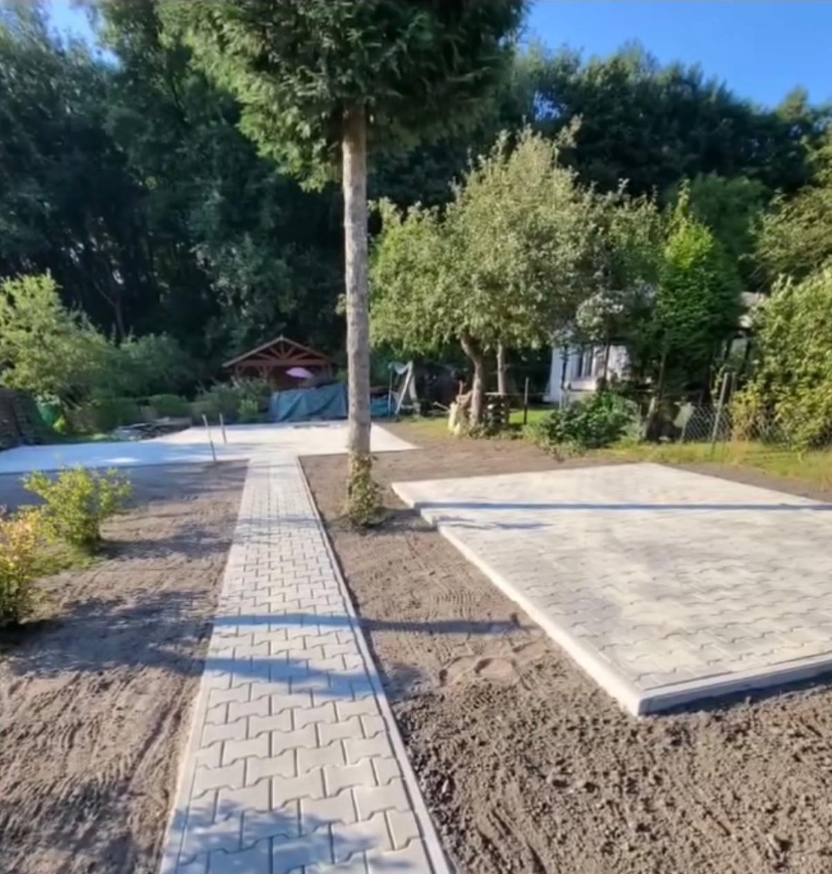 Ścieżka i plac z jasnoszarej kostki brukowej w ogrodzie, widok na altanę i zieleń w tle, świeżo ułożona kostka, częściowo przygotowany teren.