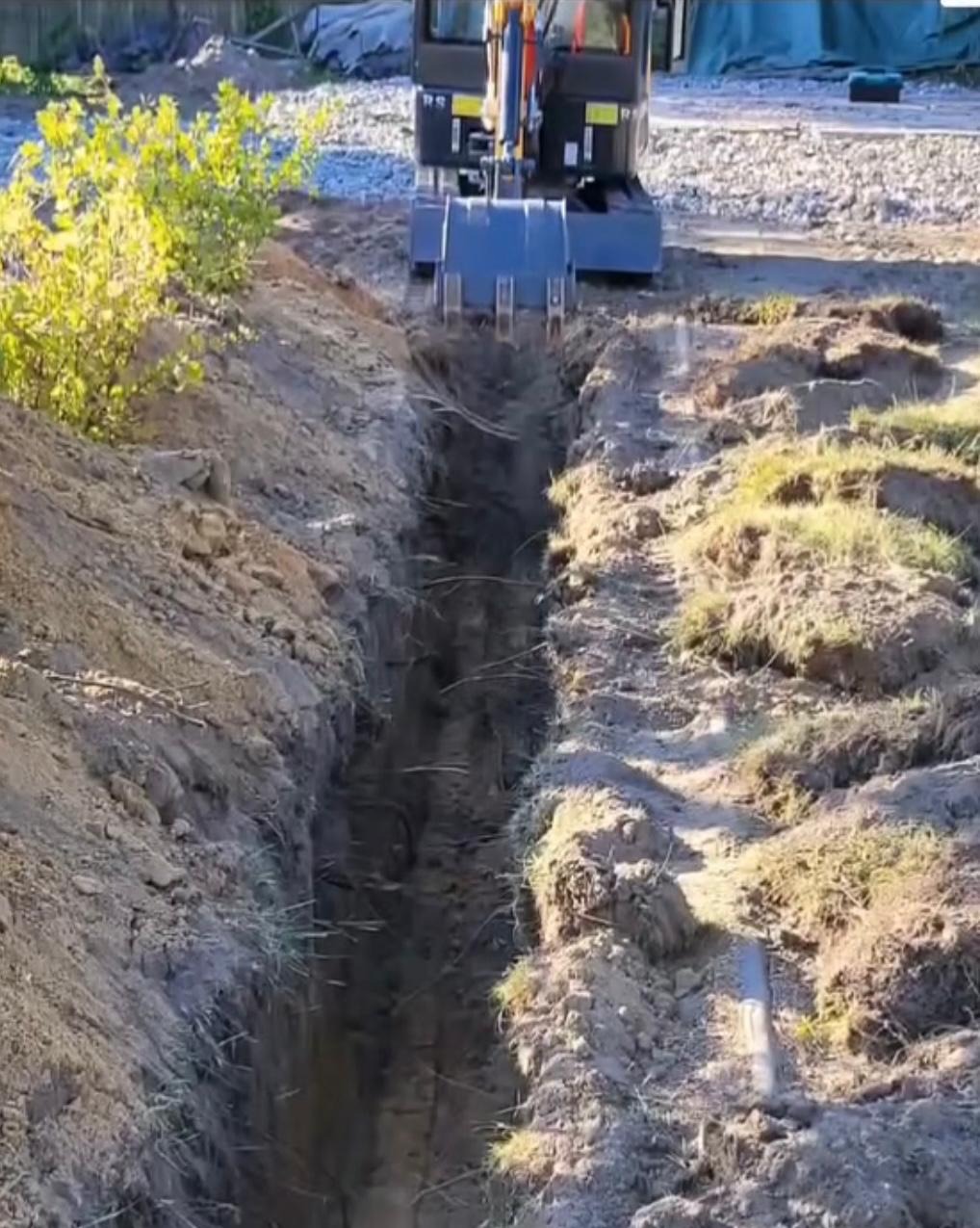 Koparka wykopuje wąski rów w ziemi. Rów biegnie w głąb kadru, a na jego końcu stoi maszyna. Tło stanowi teren budowy z widoczną roślinnością i materiałami.