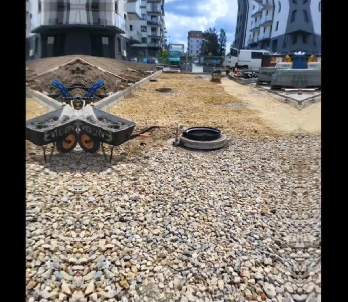 Teren przygotowany pod budowę chodnika lub drogi, z widocznym żwirem, studzienką kanalizacyjną i budynkami w tle. Widoczny wózek budowlany.