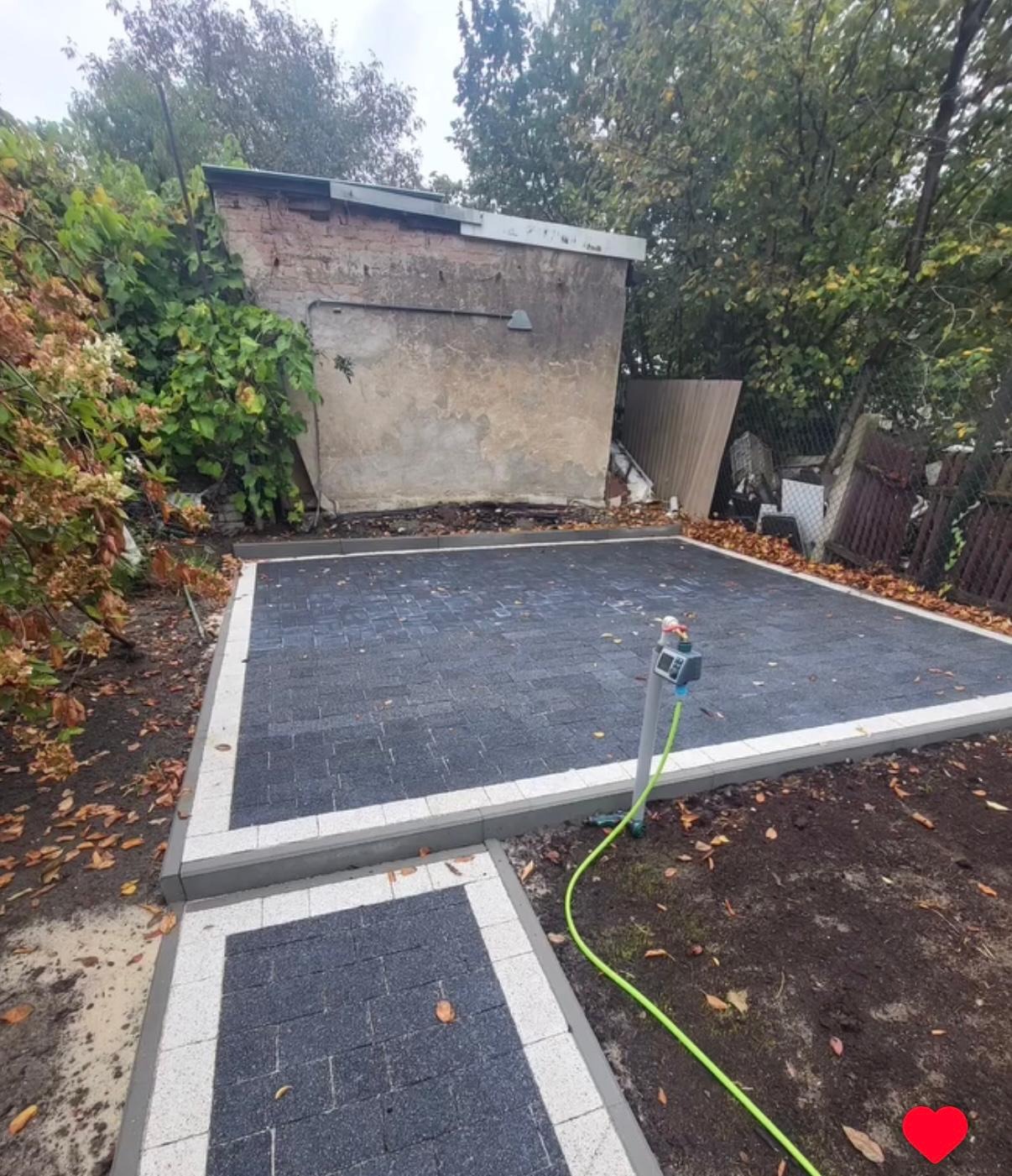 Ułożona, ciemnoszara kostka brukowa z białym obramowaniem, tworząca plac. W tle budynek gospodarczy i zieleń. Widoczny kran z podłączonym wężem ogrodowym.