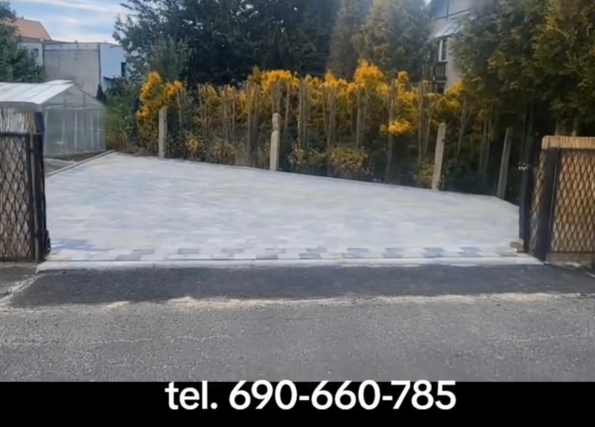 Jasnoszara kostka brukowa tworząca podjazd, widoczna przez otwartą bramę, z roślinnością w tle. Estetyczne wykończenie posesji.