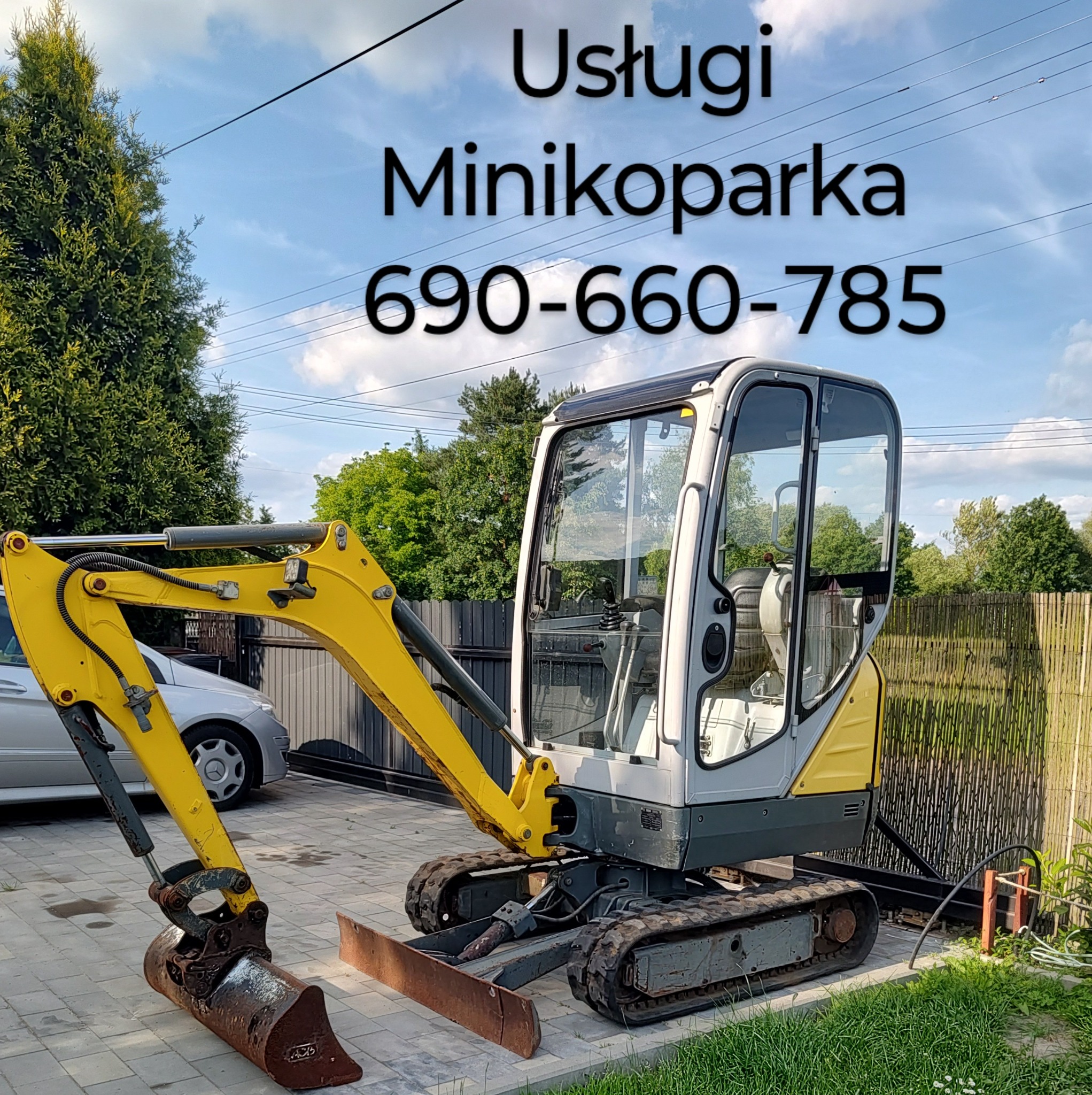 Żółto-szara minikoparka gąsienicowa na kostce brukowej, z widocznym ramieniem i łyżką, na tle zieleni i ogrodzenia. Na górze napis informacyjny z numerem telefonu.