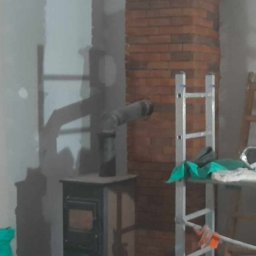 Paweł Laskowski Usługi Kominiarskie - Wnętrze w trakcie remontu z piecem typu koza podłączonym do komina obudowanego czerwoną cegłą, widoczna drabina, resztki materiałów budowlanych i worek zaprawy.