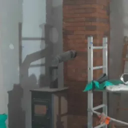 Wnętrze w trakcie remontu z piecem typu koza podłączonym do komina obudowanego czerwoną cegłą, widoczna drabina, resztki materiałów budowlanych i worek zaprawy.