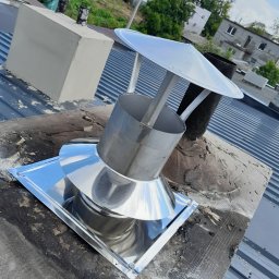 Paweł Laskowski Usługi Kominiarskie - Błyszcząca, metalowa nasada kominowa z daszkiem na kominie z szarej blachy trapezowej, w tle budynki mieszkalne i zieleń.