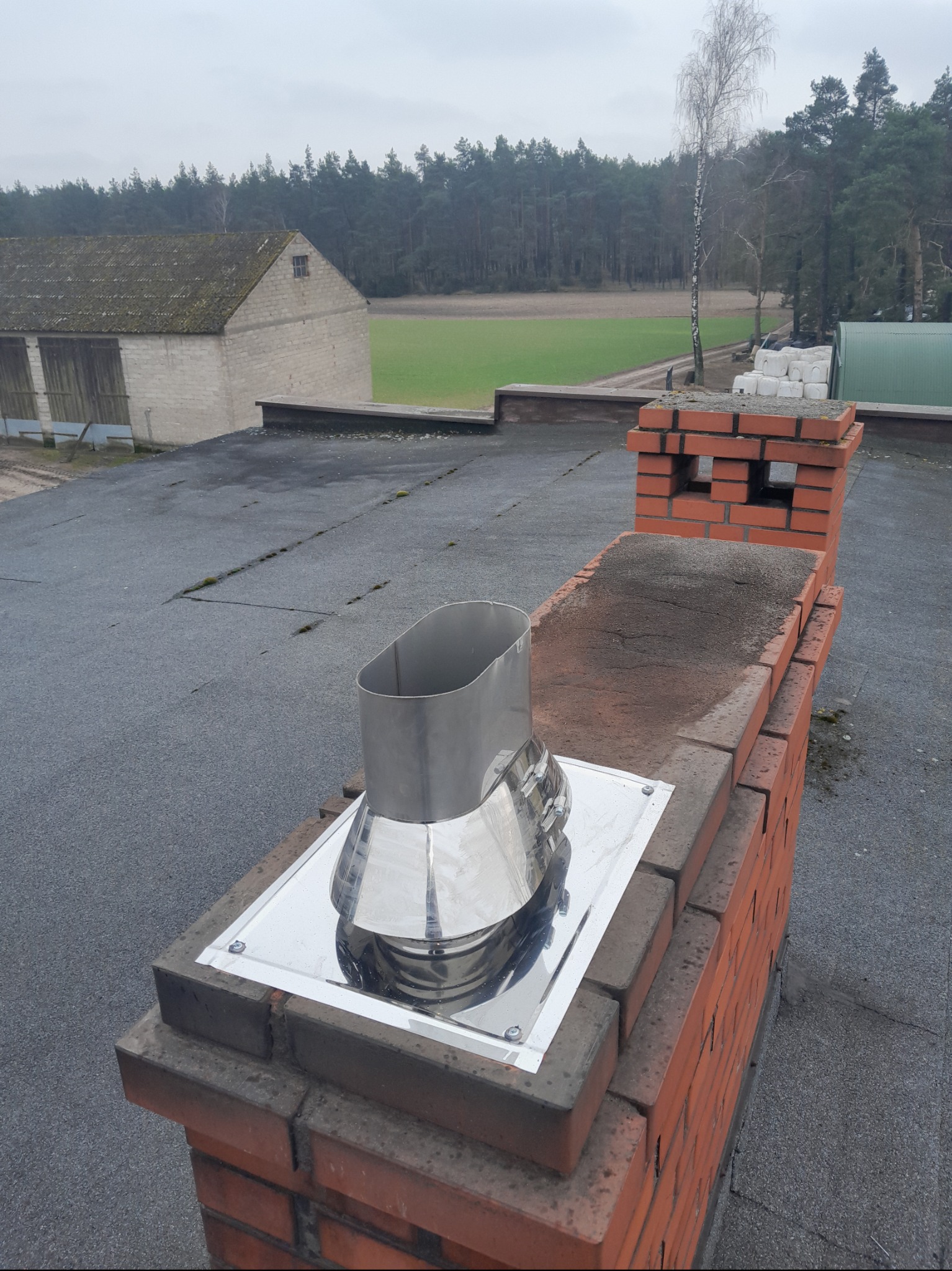 Widok z góry na komin z cegły z zamontowanym, błyszczącym, metalowym wkładem kominowym na dachu pokrytym papą. W tle budynek gospodarczy, pole i las.