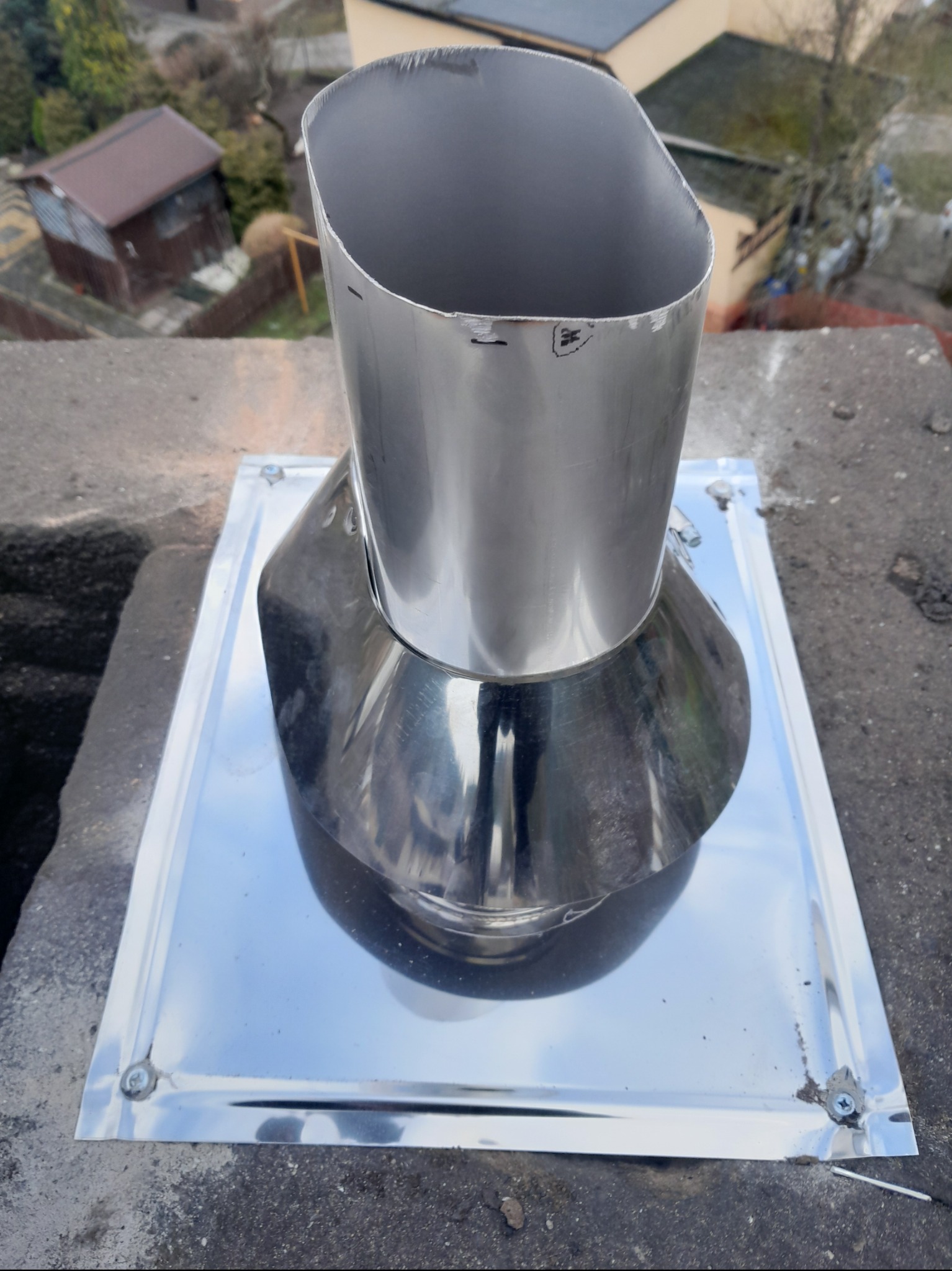 Nowa, błyszcząca, metalowa nasada kominowa z kwadratową podstawą, zamontowana na kominie z widocznym otoczeniem.