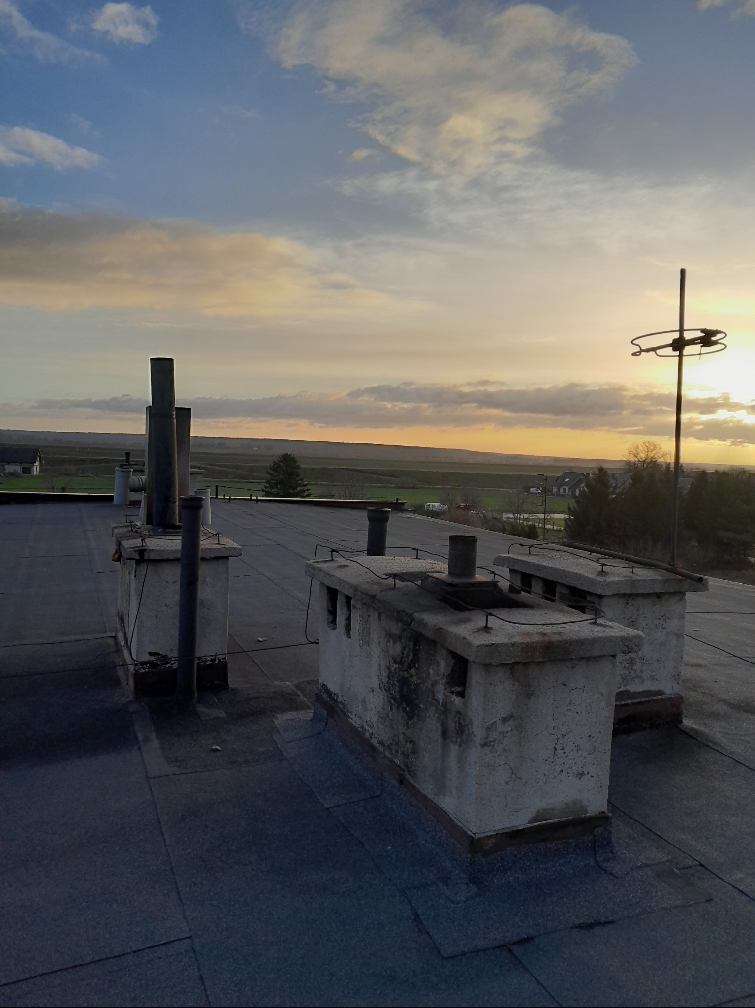Widok z dachu na kominy i antenę telewizyjną na tle zachodzącego słońca, widoczny krajobraz wiejski.