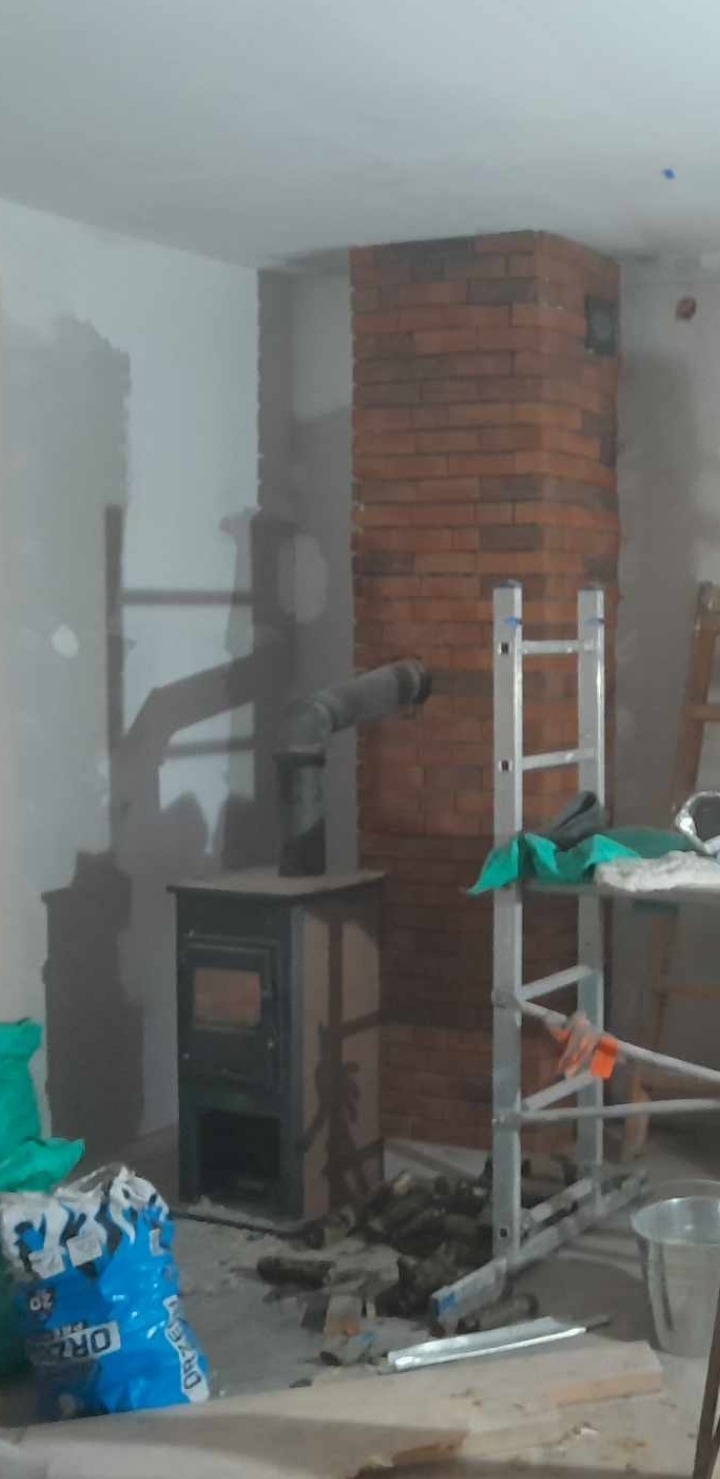 Wnętrze w trakcie remontu z piecem typu koza podłączonym do komina obudowanego czerwoną cegłą, widoczna drabina, resztki materiałów budowlanych i worek zaprawy.