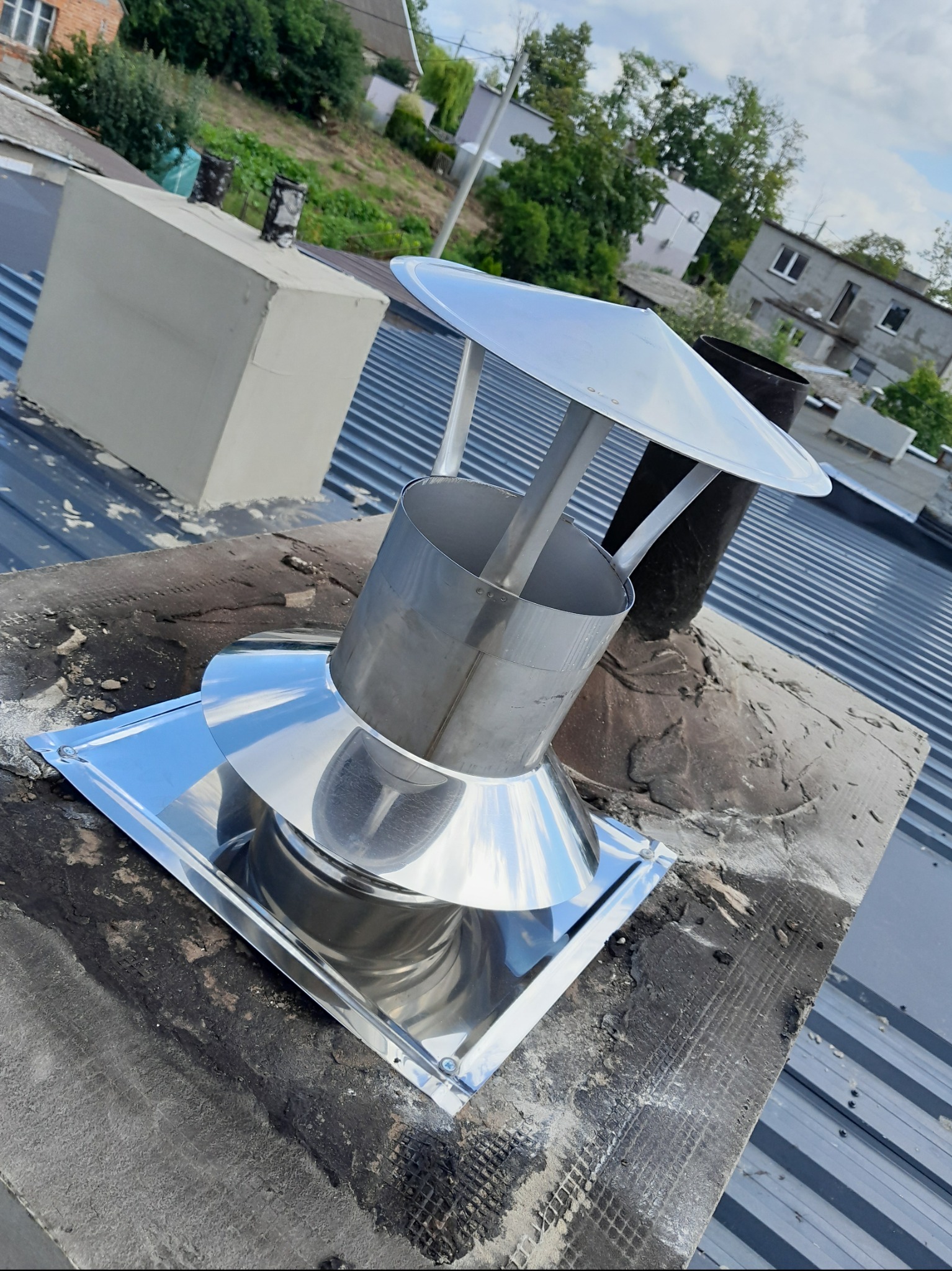 Błyszcząca, metalowa nasada kominowa z daszkiem na kominie z szarej blachy trapezowej, w tle budynki mieszkalne i zieleń.