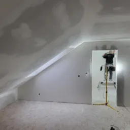 Poddasze po montażu płyt, widoczny pracownik na drabinie szpachlujący łączenia, niedokończone wykończenie, na podłodze płyty OSB i odkurzacz przemysłowy.