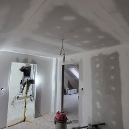 MBIT - Poddasze w trakcie wykańczania: ściany i sufit pokryte szpachlą, widoczny pracownik na drabinie, lampa halogenowa oświetla pomieszczenie, surowy stan wnętrza z widocznymi przewodami elektrycznymi.
