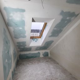 MBIT - Poddasze w trakcie prac wykończeniowych: ściany przygotowane do szpachlowania, widoczne łączenia płyt, okno dachowe z widokiem na budynek, wystające przewody elektryczne, narzędzia na podłodze.