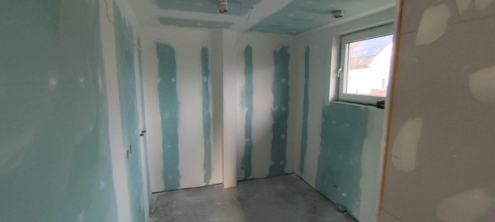 Wnętrze w trakcie prac wykończeniowych: ściany i sufit pokryte płytami, widoczne łączenia płyt wymagające szpachlowania, surowy beton na podłodze, okno z widokiem na dom.