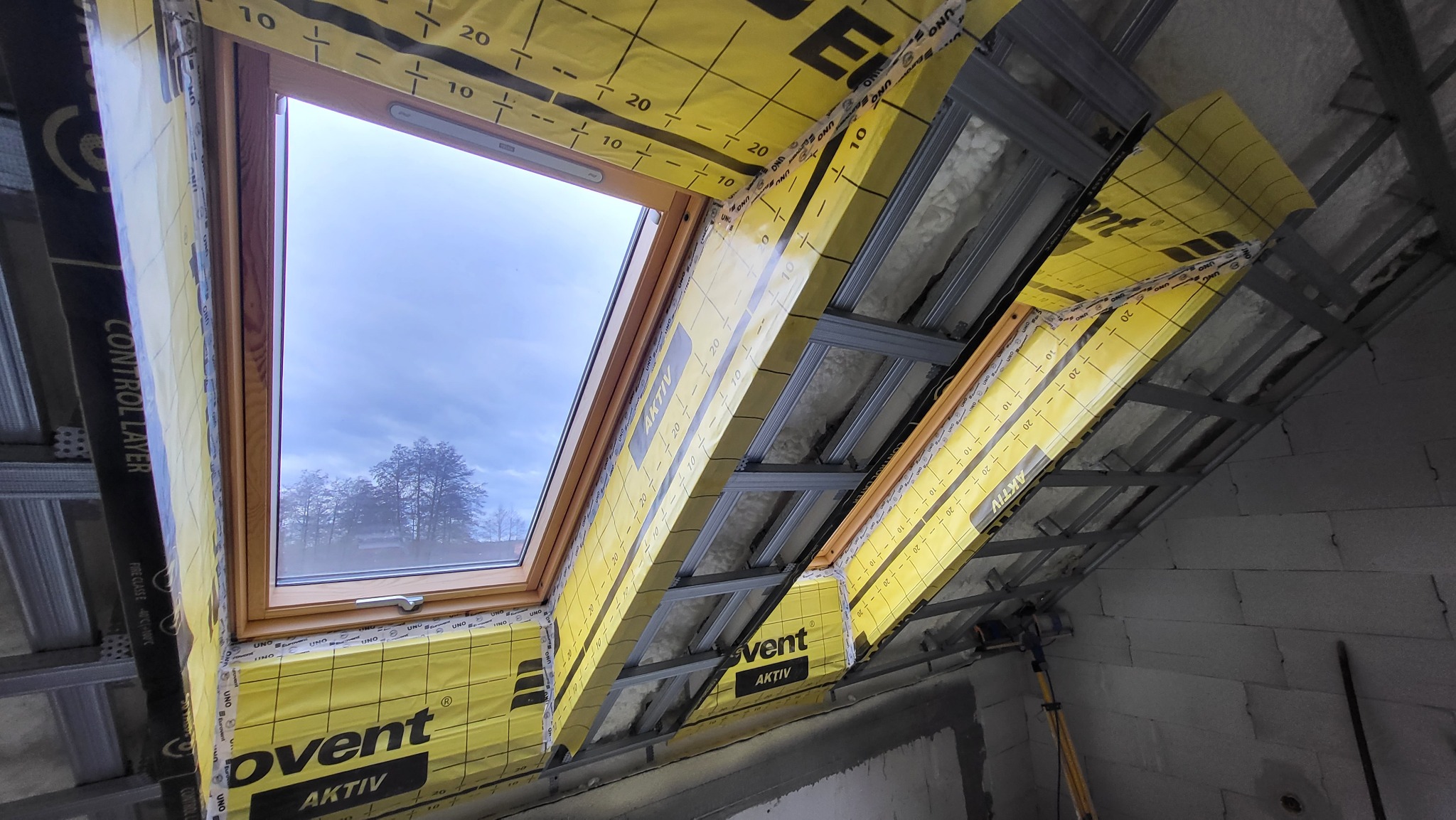 Montaż okna dachowego z widoczną konstrukcją metalową, izolacją i folią paroizolacyjną z nadrukiem, w tle widok na drzewa przez szybę.