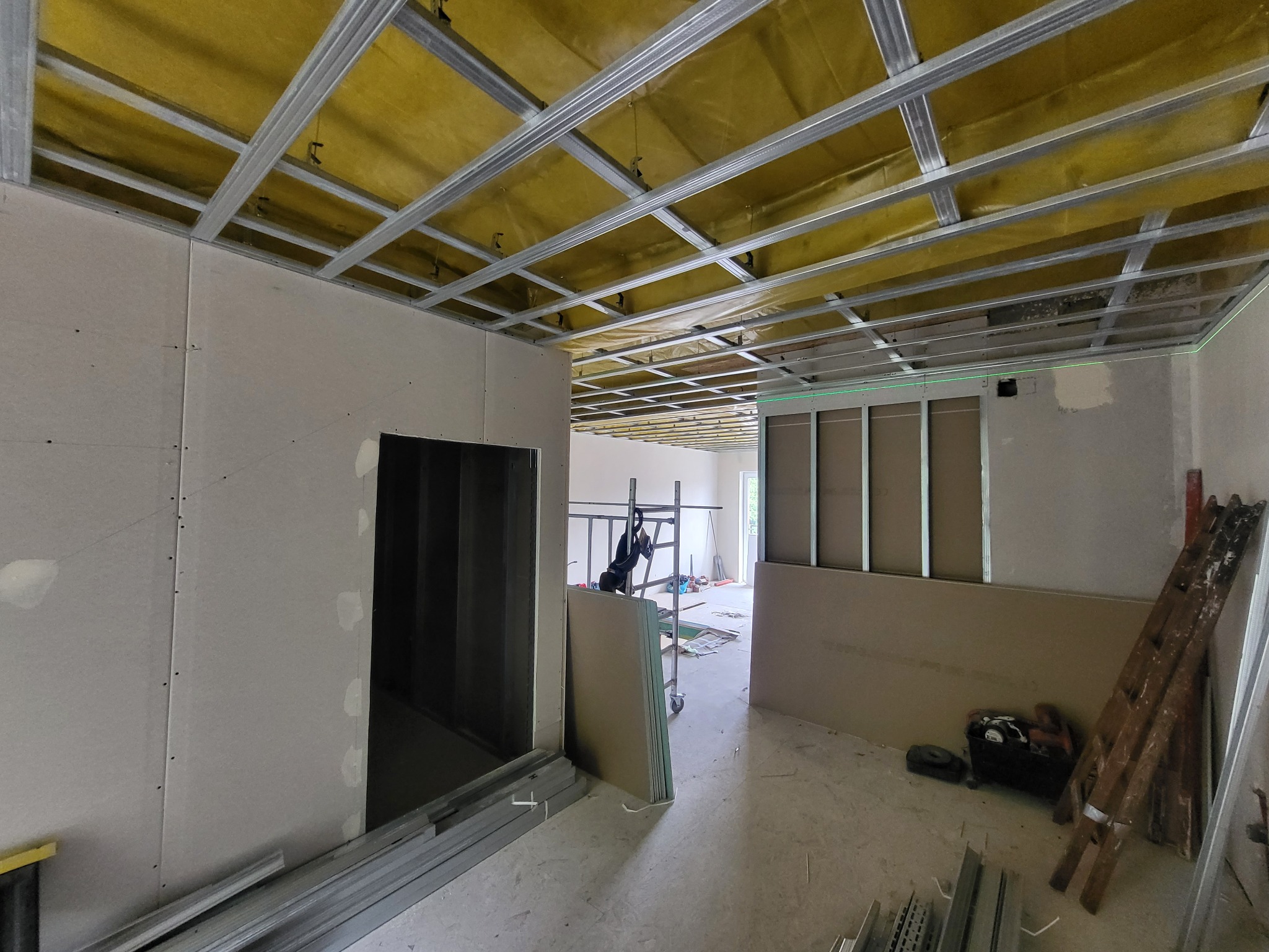 Montaż stelaża sufitowego z profili metalowych, ocieplenie wełną mineralną widoczne w przestrzeni między profilami, częściowo obłożone płytami gipsowo-kartonowymi, widoczna drabina i narzędzia.