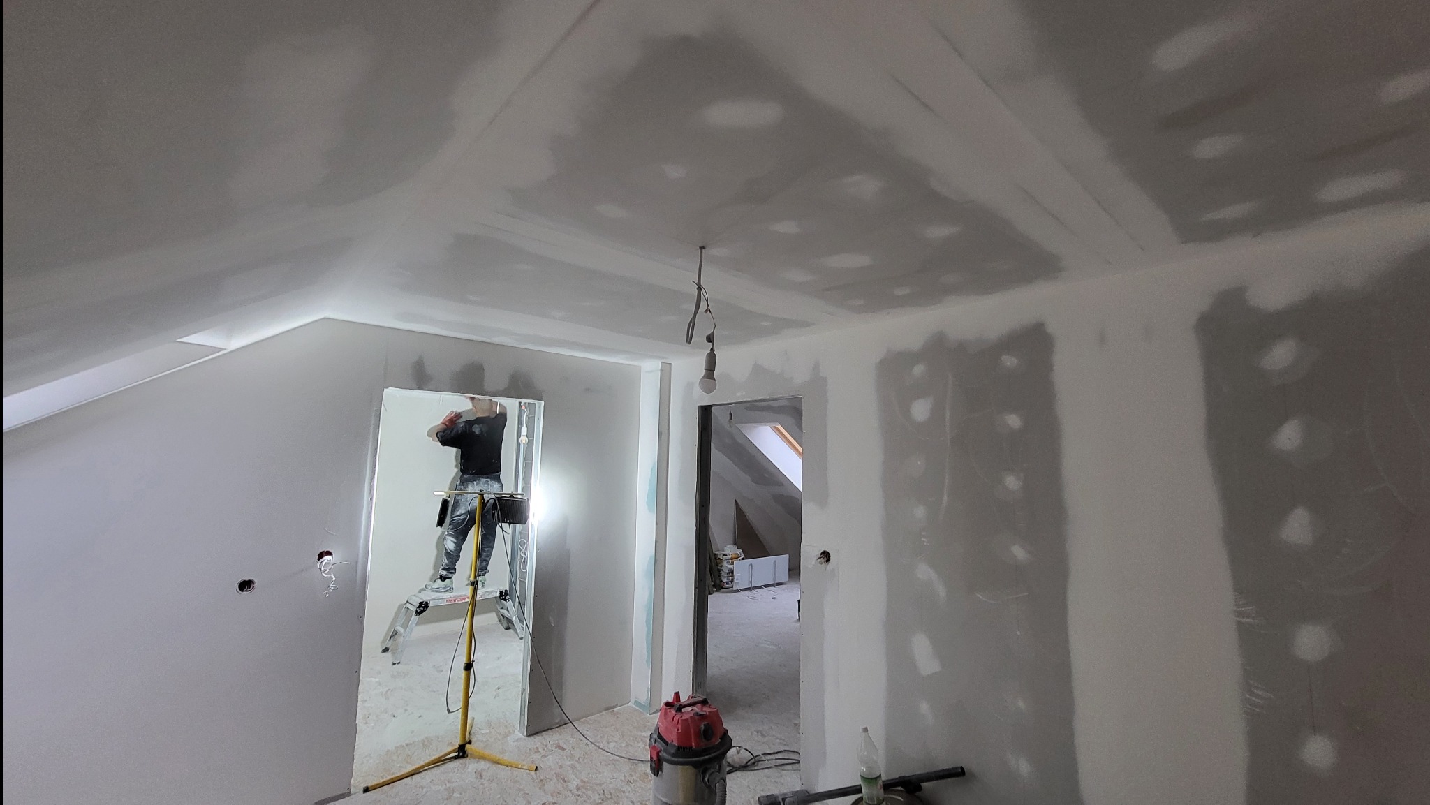 Poddasze w trakcie wykańczania: ściany i sufit pokryte szpachlą, widoczny pracownik na drabinie, lampa halogenowa oświetla pomieszczenie, surowy stan wnętrza z widocznymi przewodami elektrycznymi.