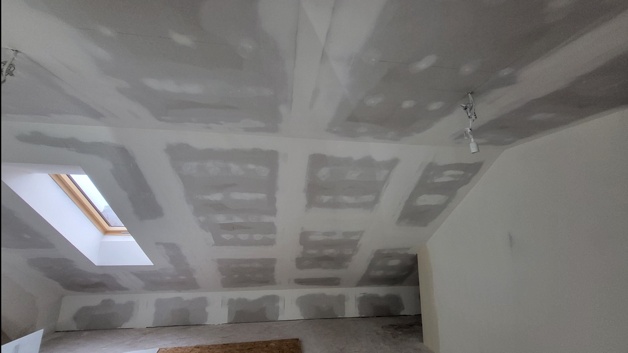 Poddasze w trakcie prac wykończeniowych: widoczne płyty na suficie i ścianach, częściowo zaszpachlowane łączenia, okno dachowe i prowizoryczne oświetlenie.