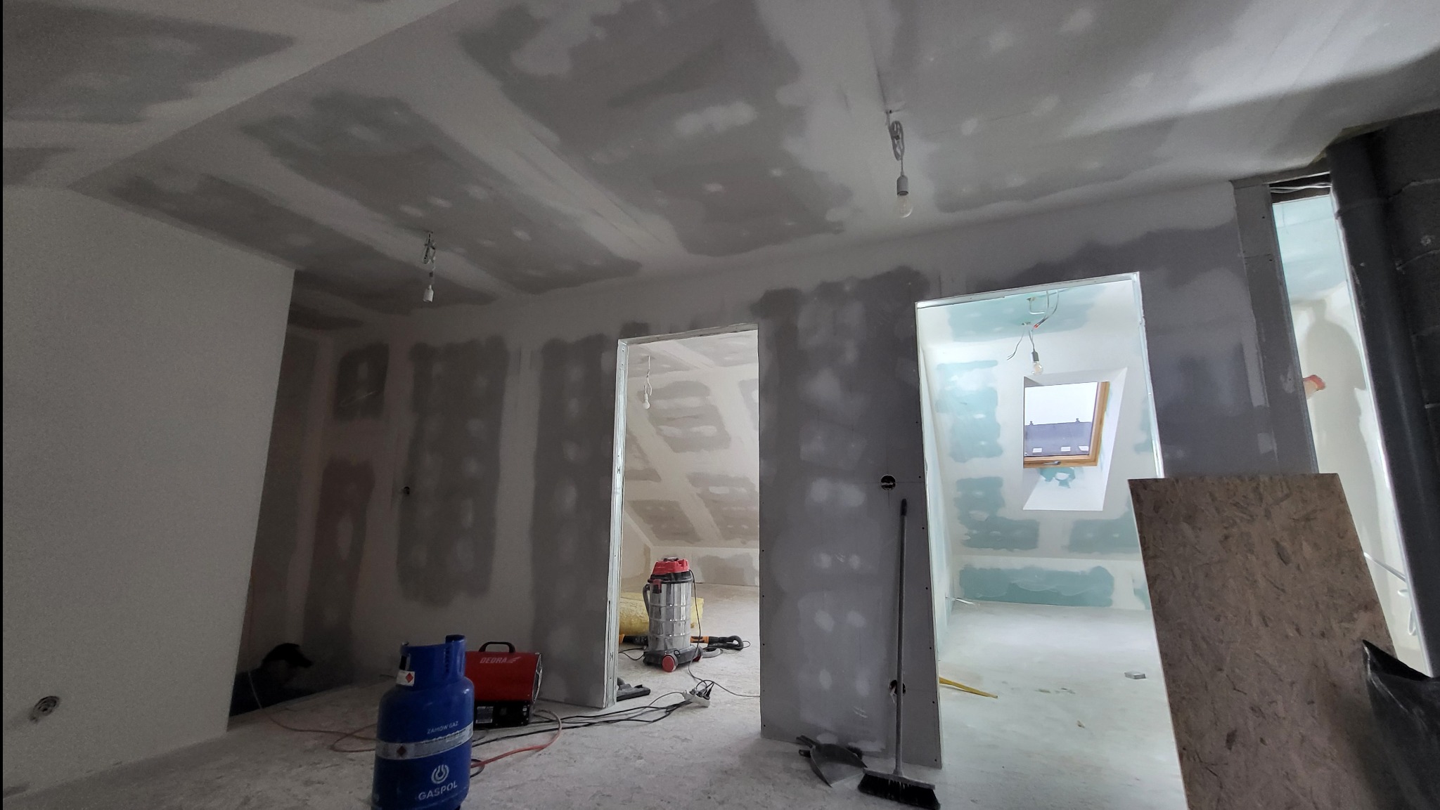 Wnętrze w trakcie prac wykończeniowych: ściany i sufit pokryte płytami, widoczne szpachlowanie, butla gazowa, odkurzacz przemysłowy i narzędzia.