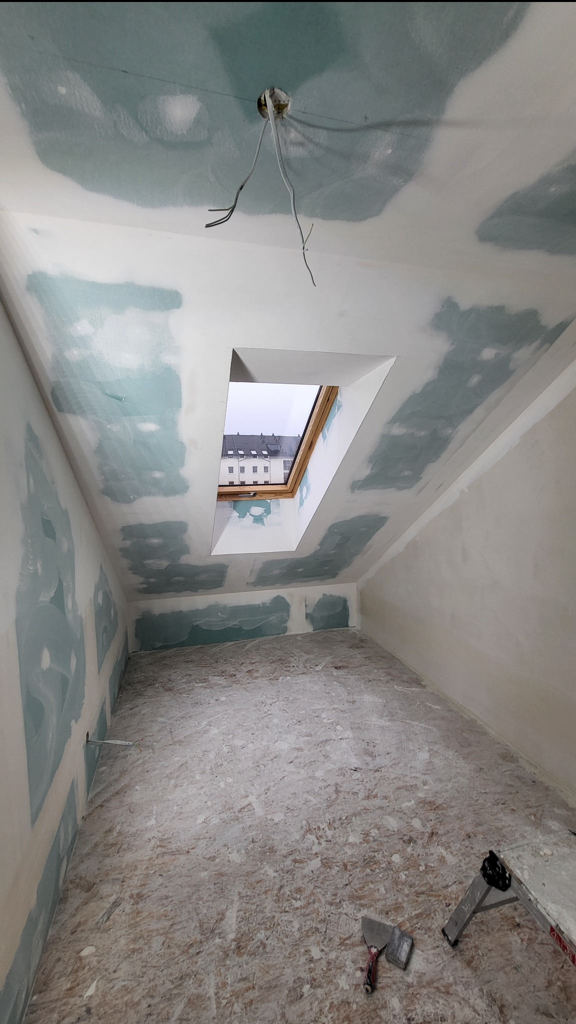 Poddasze w trakcie prac wykończeniowych: ściany przygotowane do szpachlowania, widoczne łączenia płyt, okno dachowe z widokiem na budynek, wystające przewody elektryczne, narzędzia na podłodze.