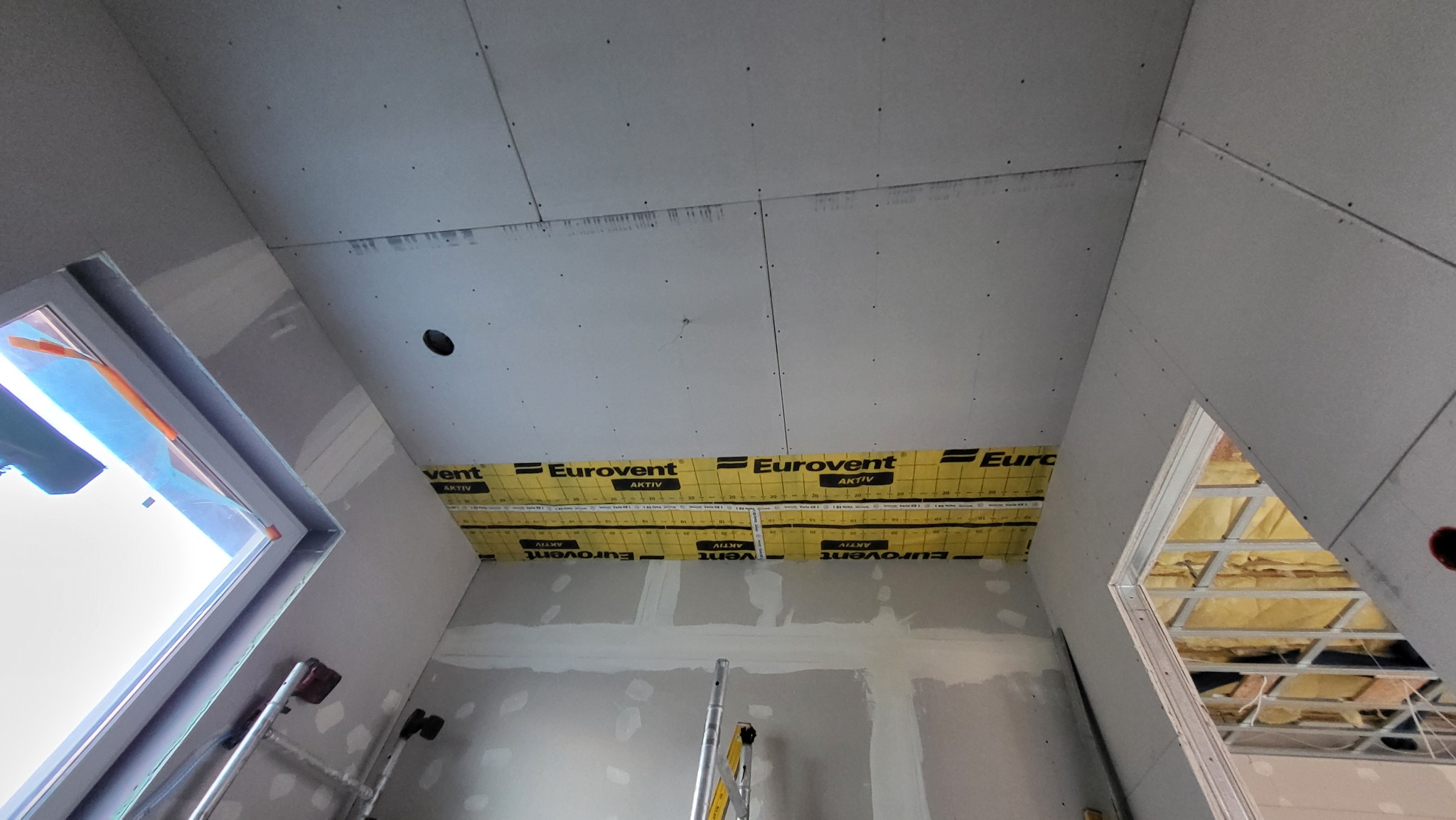 Wnętrze w trakcie prac wykończeniowych: zamontowane płyty na suficie, widoczna izolacja Eurovent, ściany szpachlowane, drabina aluminiowa w rogu pomieszczenia.