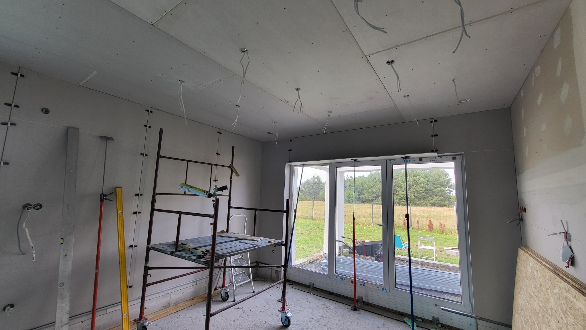 Wnętrze w trakcie montażu sufitu podwieszanego z widocznymi przewodami elektrycznymi, rusztowaniem, poziomnicami i oknem z widokiem na zieleń.