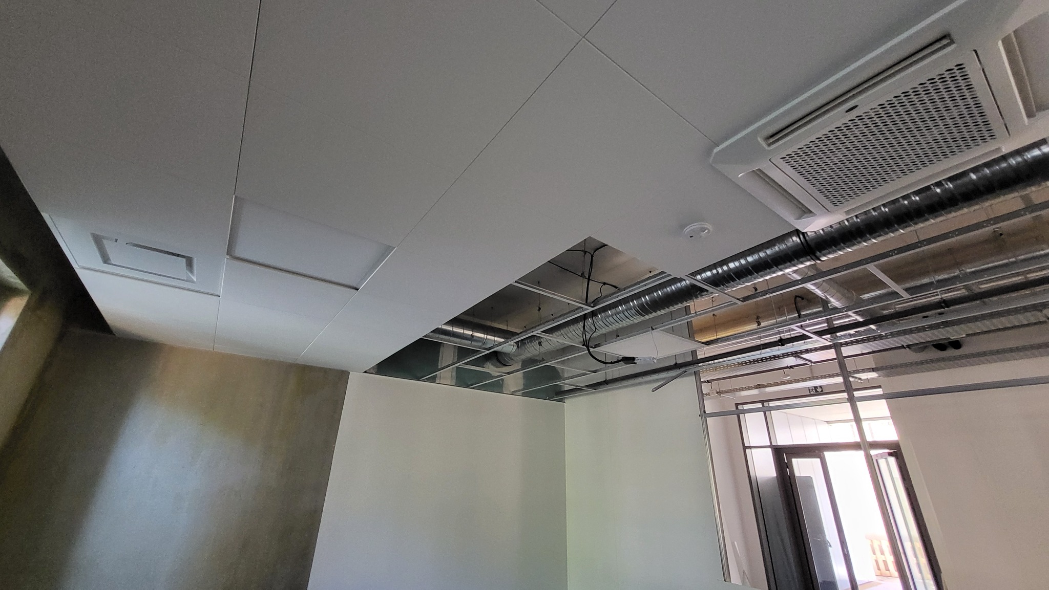 Montaż sufitu podwieszanego z płyt kartonowo-gipsowych z widoczną instalacją wentylacyjną i klimatyzacją. Częściowo wykończone ściany w jasnych barwach.