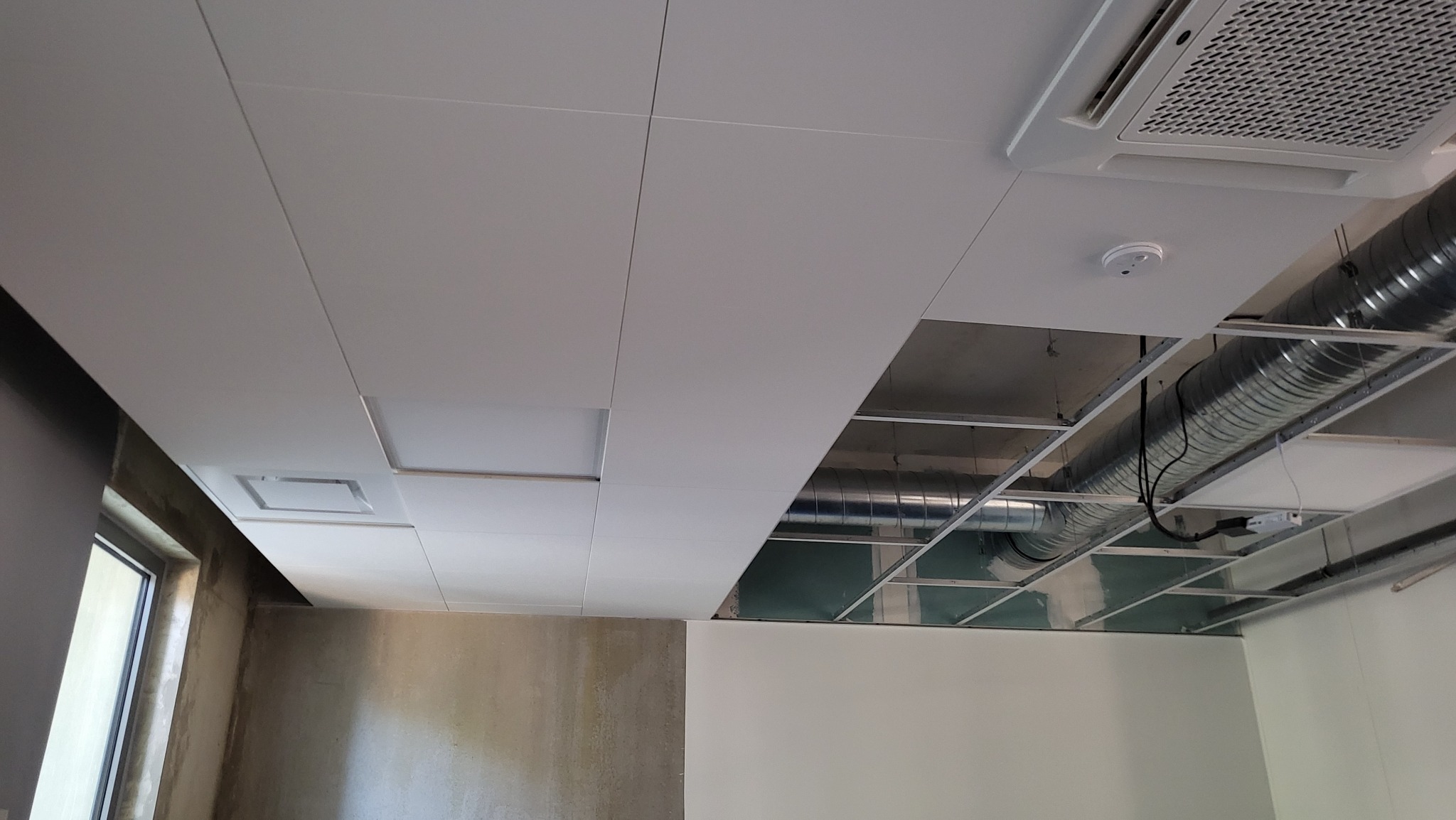 Montaż sufitu podwieszanego z widoczną instalacją wentylacyjną i klimatyzacją. Częściowo wykończony sufit z panelami i oświetleniem.
