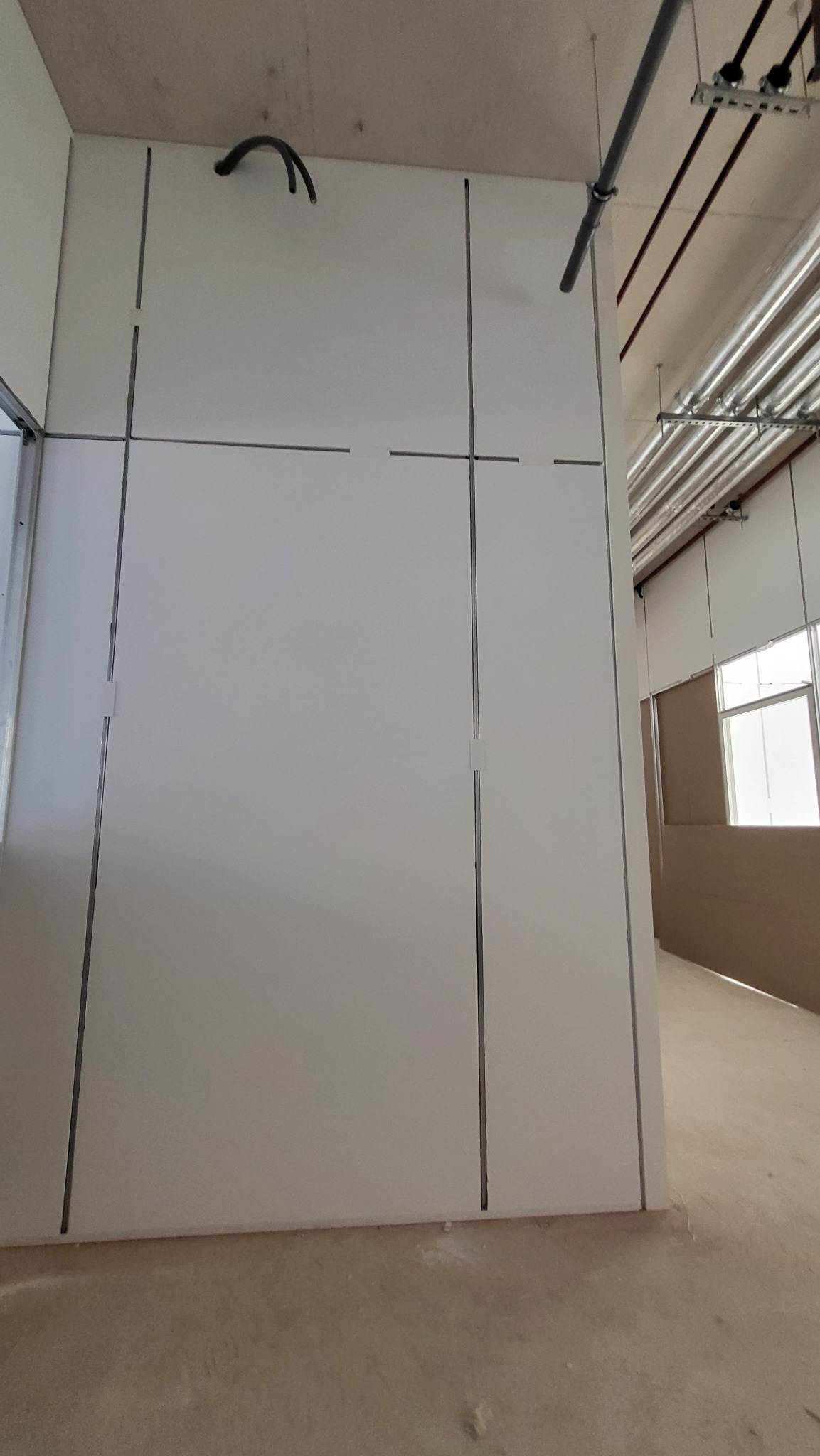 Ściana z płyt gipsowo-kartonowych w trakcie montażu, widoczne profile i łączenia. W tle instalacje pod sufitem i fragment okna. Surowy stan wykończenia podłogi.