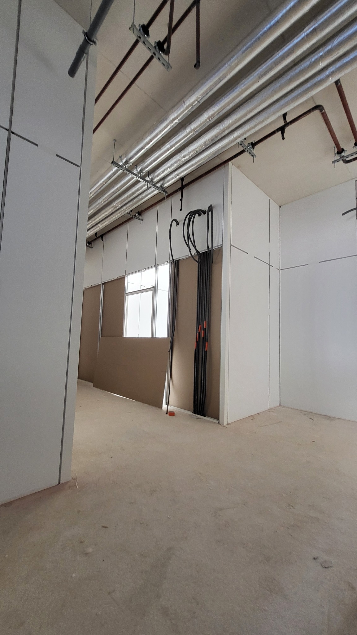 Wnętrze w trakcie prac wykończeniowych: białe ściany z płyt, widoczne instalacje pod sufitem i kable w ścianie. Surowy stan podłogi, okno w tle.