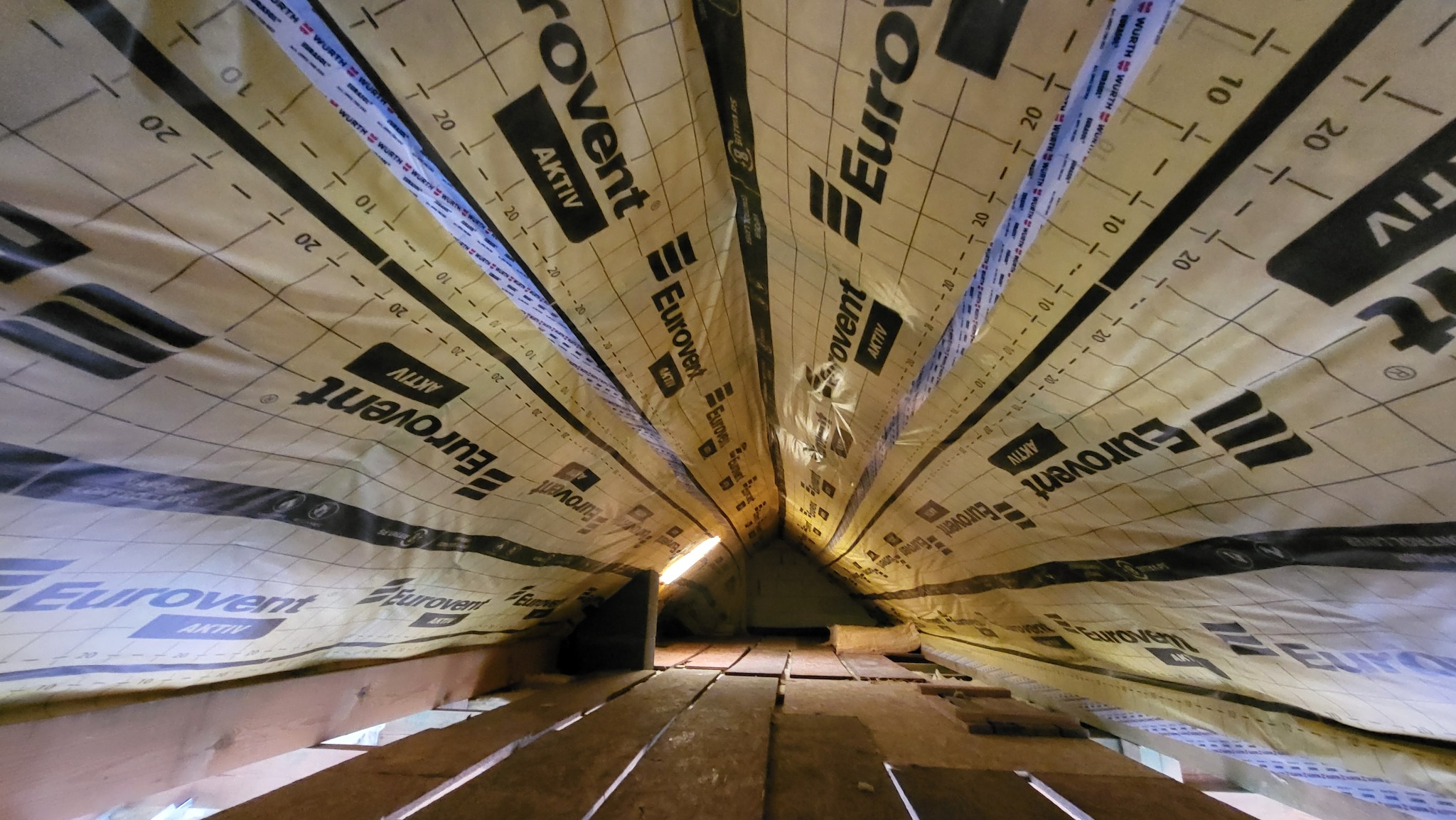 Montaż folii paroizolacyjnej Eurovent Aktiv na drewnianej konstrukcji dachu, widok z dołu. Widoczne listwy i elementy mocujące. Oświetlenie punktowe w tle.