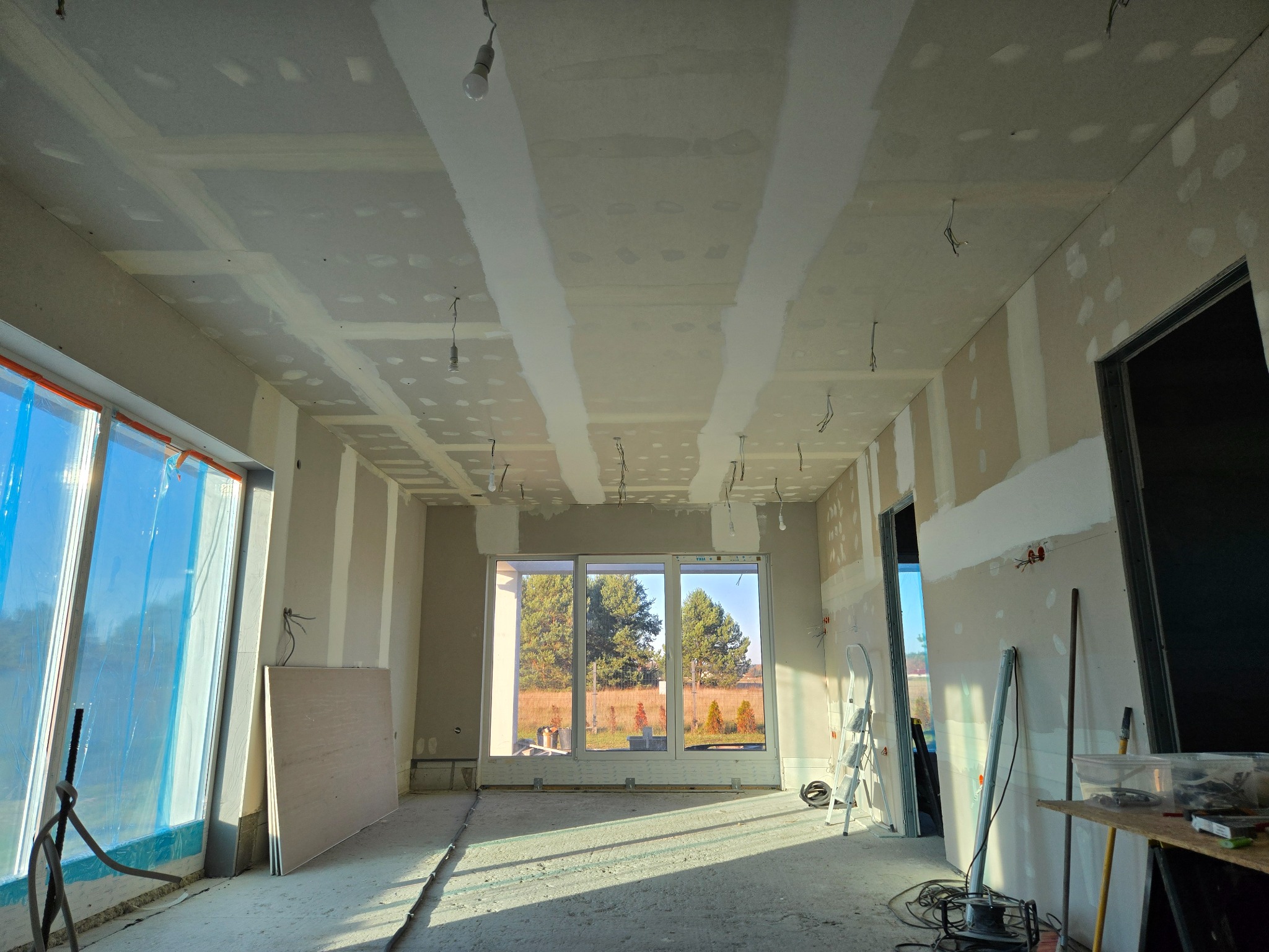 Wnętrze w trakcie prac wykończeniowych: ściany z płyt gipsowo-kartonowych, częściowo wyszpachlowane, widoczne okna z widokiem na zewnątrz, narzędzia pracy.