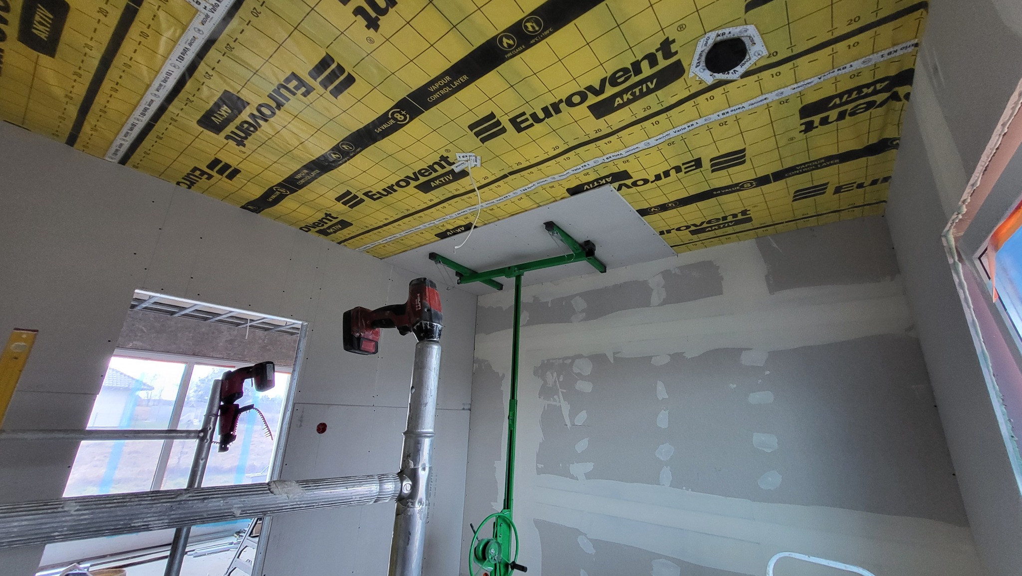 Montaż płyty karton-gips na suficie z użyciem podnośnika i wkrętarki. Widoczna folia paroizolacyjna Eurovent i szpachlowane ściany. Praca w toku.