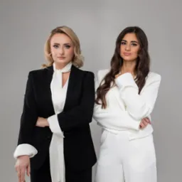 Portret dwóch kobiet w eleganckich garniturach na szarym tle: blondynka w czarnym garniturze z białą apaszką i brunetka w białym garniturze.