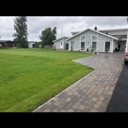 CKkoparki - Ułożona kostka brukowa prowadząca do nowoczesnego domu z szarą elewacją i dużym trawnikiem w pochmurny dzień.