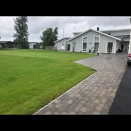 Ułożona kostka brukowa prowadząca do nowoczesnego domu z szarą elewacją i dużym trawnikiem w pochmurny dzień.