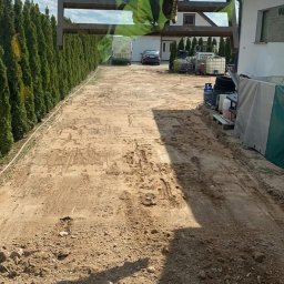 CKkoparki - Przygotowanie podłoża pod kostkę brukową z użyciem walca wibracyjnego zawieszonego na ramieniu koparki, widoczne ślady opon, obok rząd tuj i budynek.