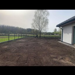 CKkoparki - Przygotowanie terenu pod trawnik: wyrównana, ciemna ziemia, widoczny pracownik z grabiami pod samotnym drzewem brzozy, w tle ogrodzenie panelowe i zieleń.