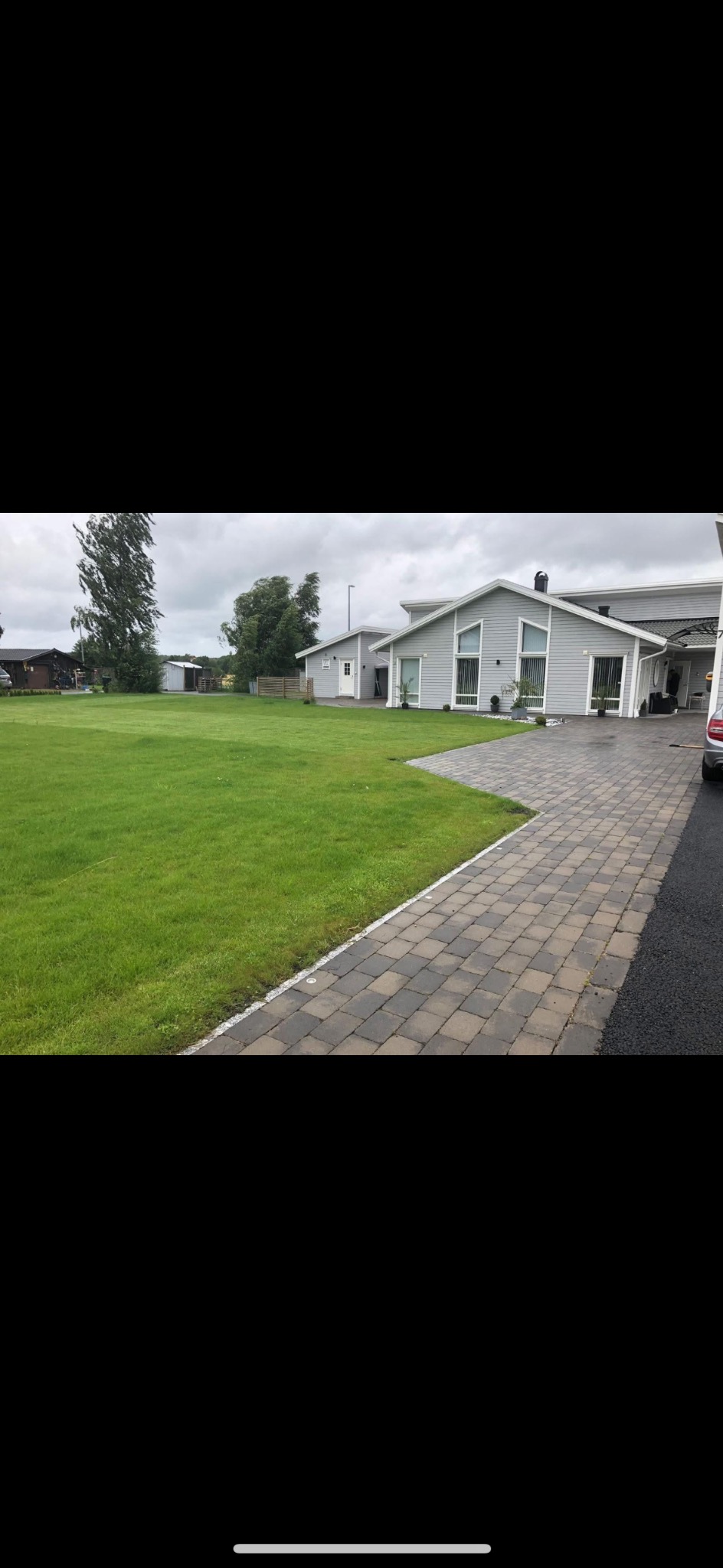 Ułożona kostka brukowa prowadząca do nowoczesnego domu z szarą elewacją i dużym trawnikiem w pochmurny dzień.