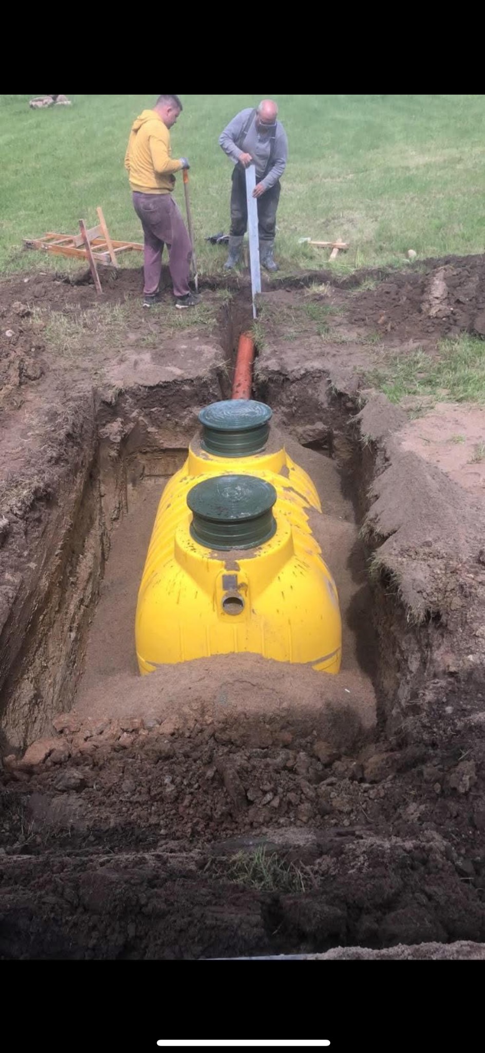 Żółty zbiornik na szambo umieszczony w wykopie, częściowo zasypany piaskiem, z widoczną rurą odpływową i dwoma zielonymi pokrywami, dwóch mężczyzn pracuje przy wykopie, jeden z łopatą, drugi...