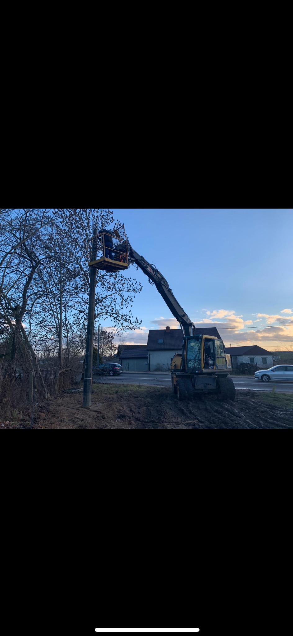 Żółta koparka z wysięgnikiem i platformą, na której stoi pracownik, przycina gałęzie drzewa przy drodze, w tle widoczny budynek i przejeżdżające samochody.
