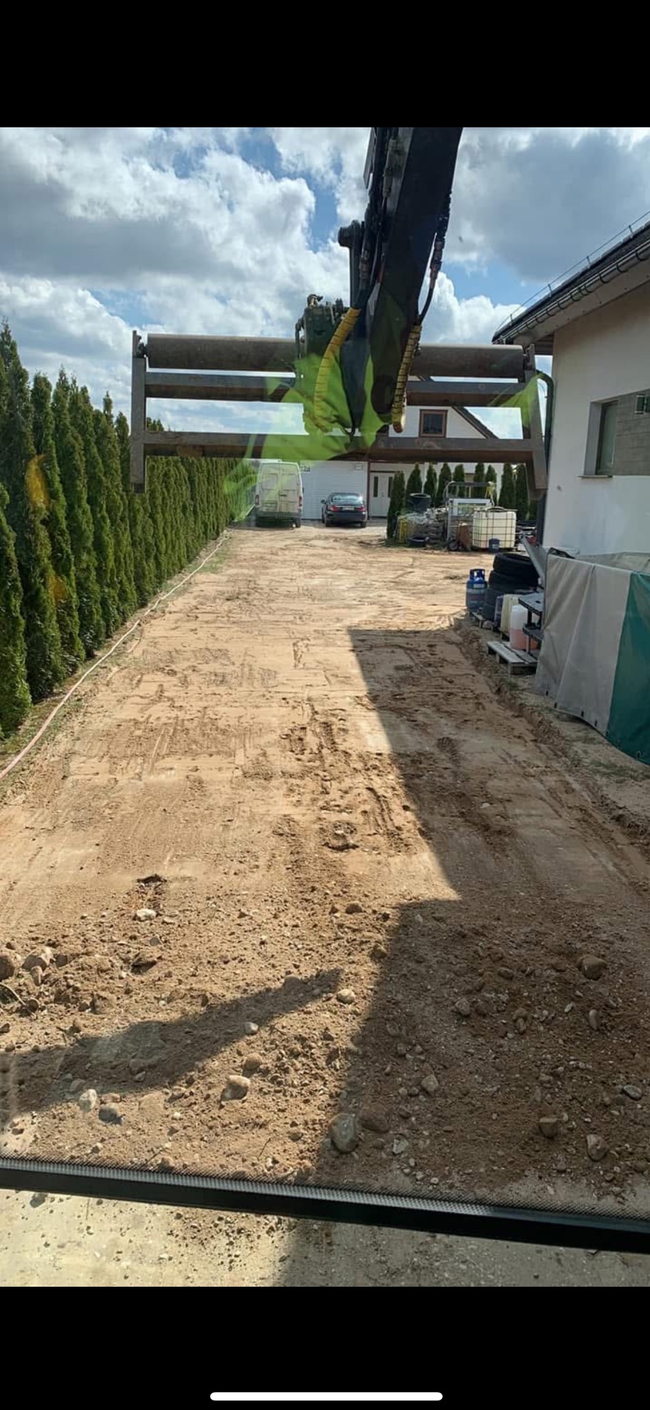 Przygotowanie podłoża pod kostkę brukową z użyciem walca wibracyjnego zawieszonego na ramieniu koparki, widoczne ślady opon, obok rząd tuj i budynek.