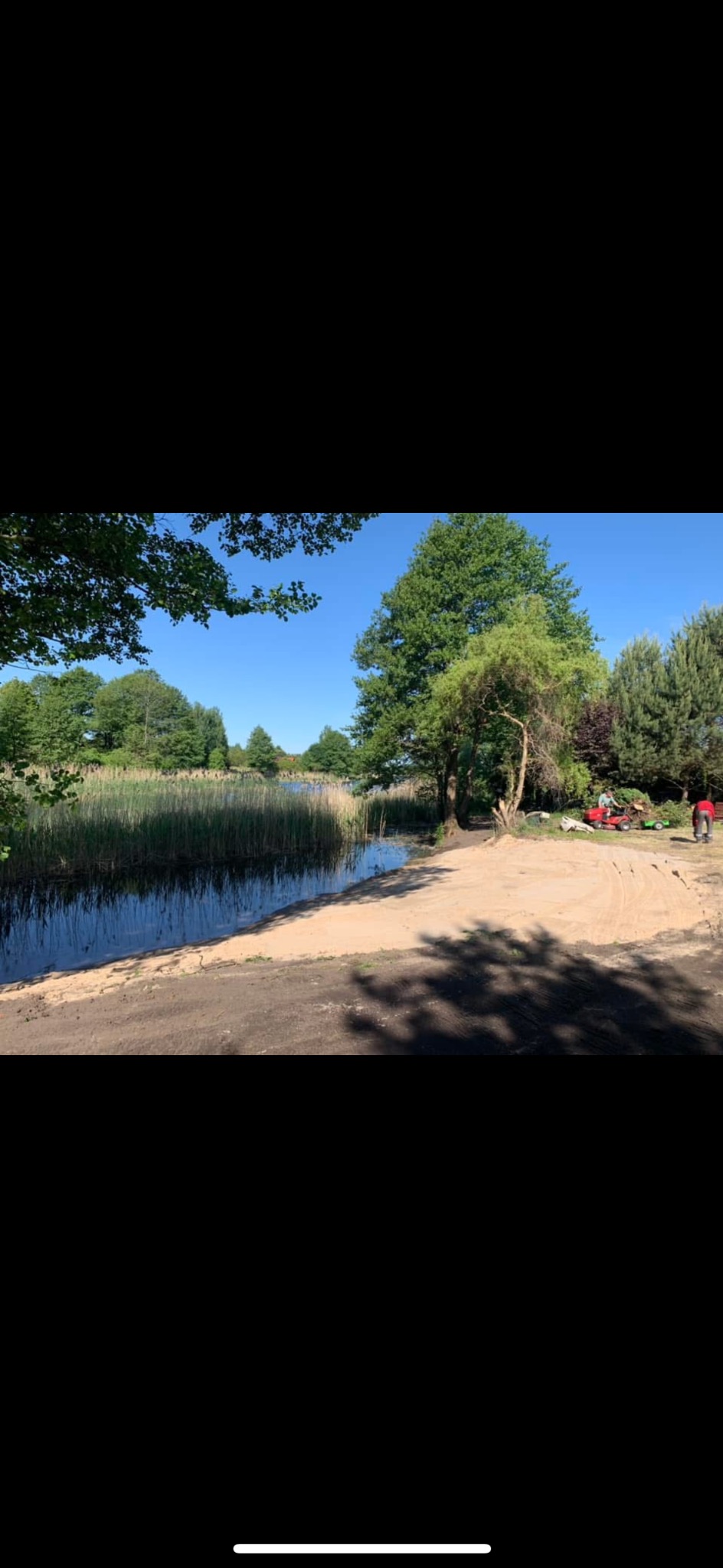 Brzeg jeziora z wyczyszczonym terenem i dwoma kosiarkami do trawy w tle, sugerujące prace porządkowe lub przygotowawcze.