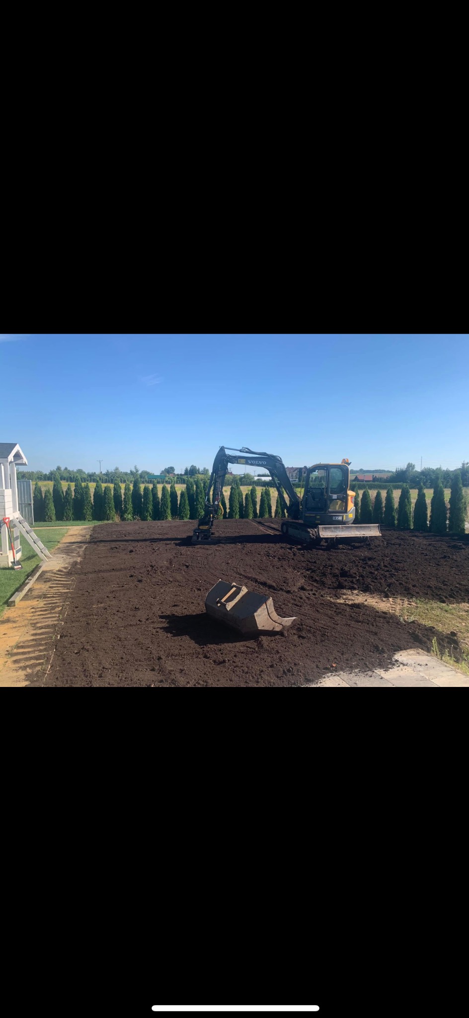 Żółto-czarna koparka Volvo EC55B na placu budowy, przygotowującym teren pod przyszłe prace, z widocznym odłożonym łyżką i rzędem tuj w tle.