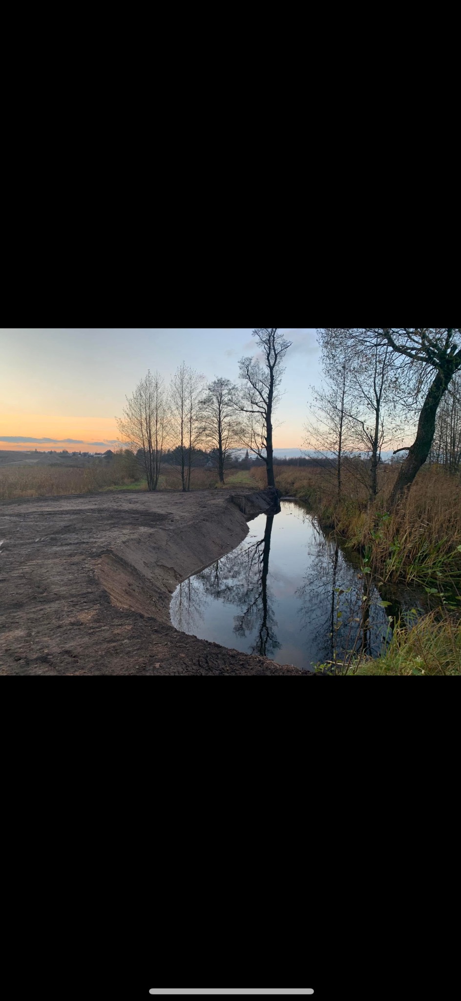 Świeżo wykopany rów melioracyjny z wodą, odbijający nagie drzewa na tle zachodzącego słońca i krajobrazu polnego.