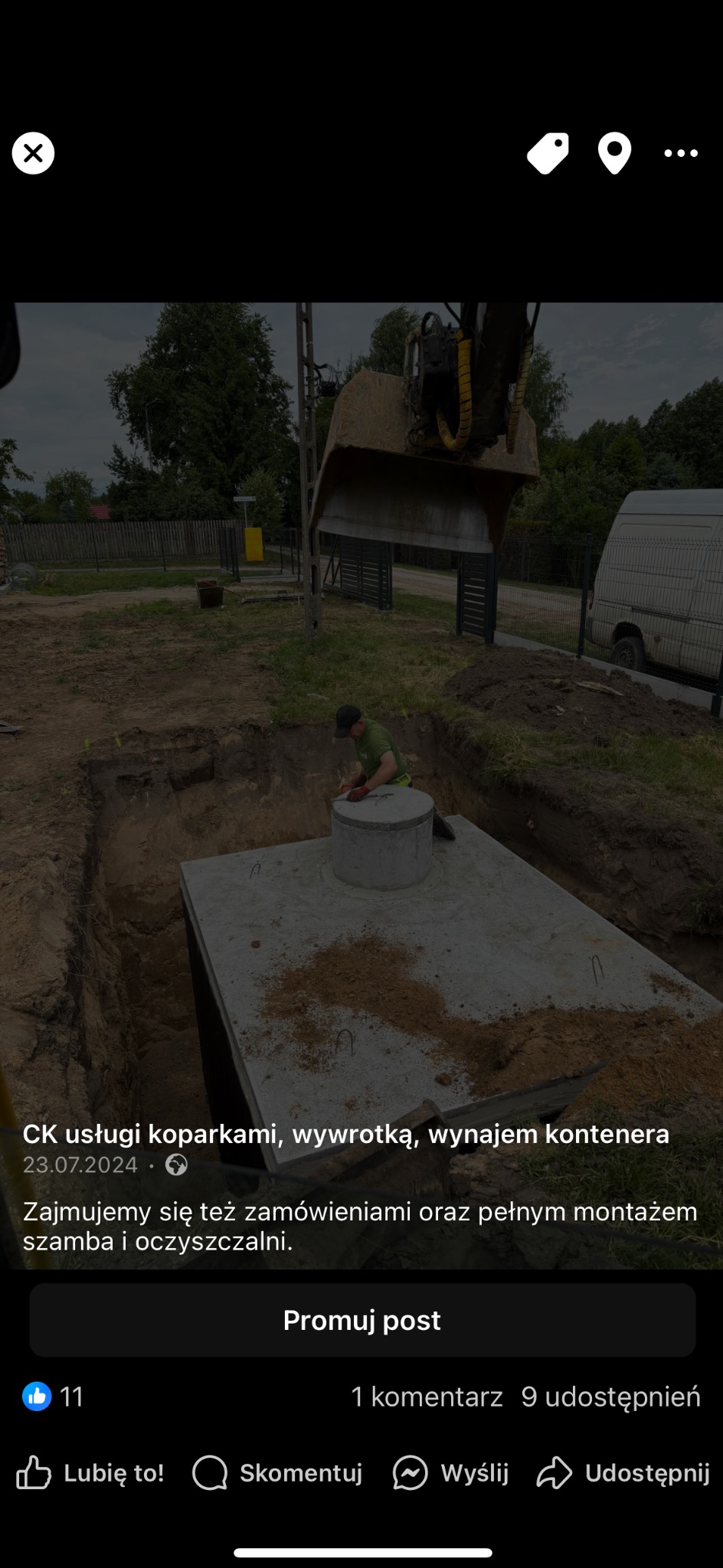 Montaż betonowego zbiornika na szambo w wykopie; pracownik w zielonej koszulce mocuje pokrywę, a w tle widoczna koparka i ogrodzenie posesji.