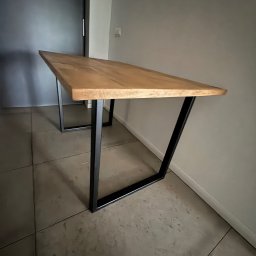 FK TECH - Stół z blatem z jasnego drewna i czarnymi, metalowymi nogami w kształcie trapezu, ustawiony na szarej podłodze z płytek.