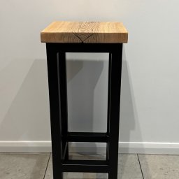 FK TECH - Drewniany taboret z blatem z jasnego drewna o widocznym usłojeniu i czarną, metalową ramą na tle białej ściany i szarej podłogi.